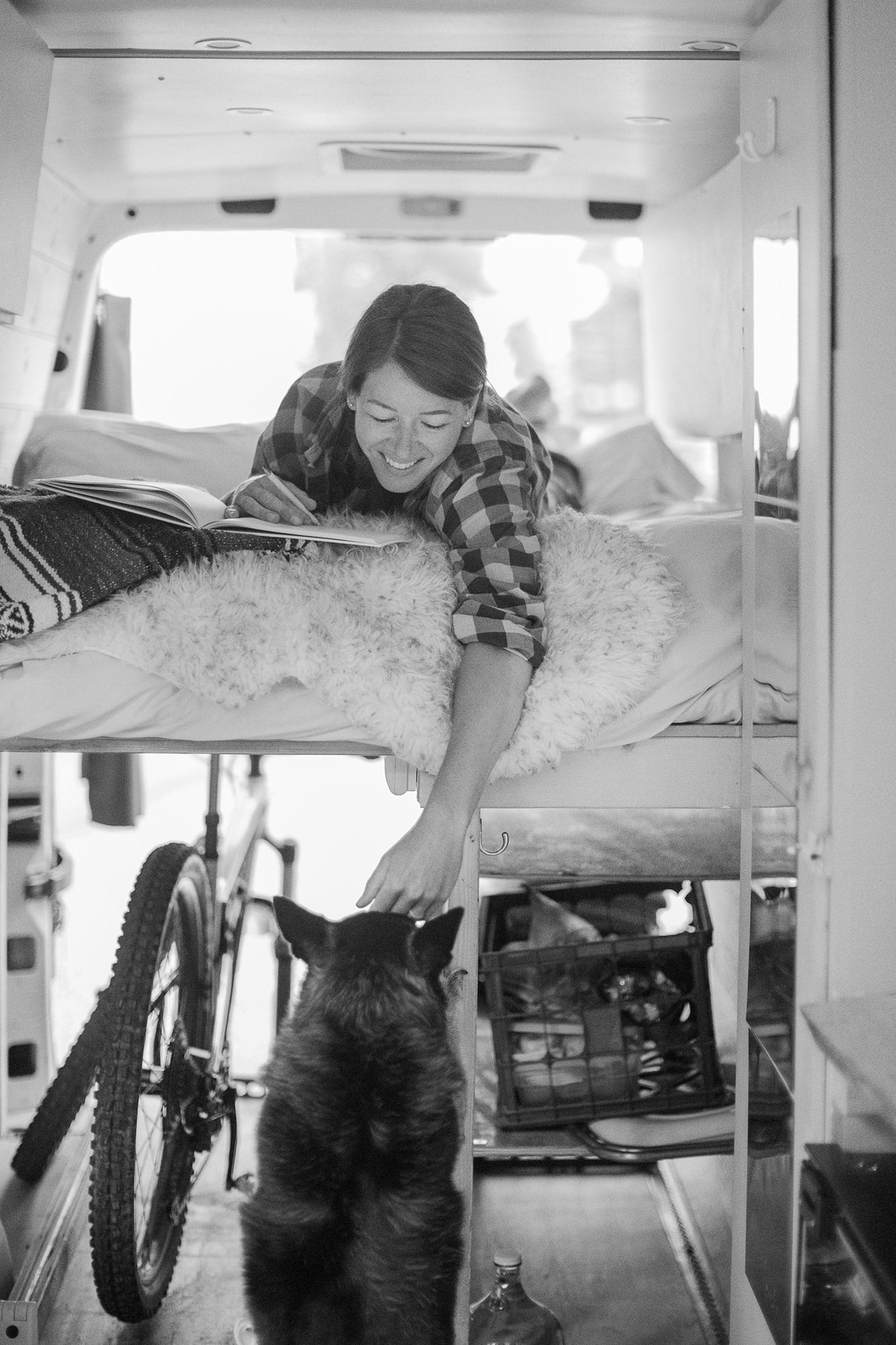 RV rental life: A smiling woman writes and pets her dog in their cozy van home, bike stored, ready for adventure.