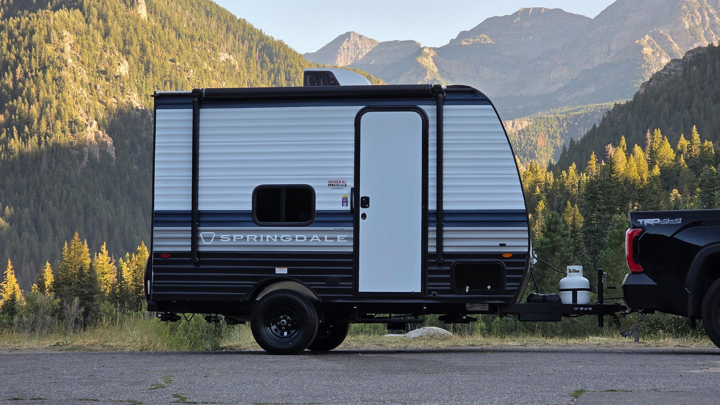 An RV rental, a Springdale travel trailer, is hitched to a truck, ready for adventure in front of majestic forested mountains.