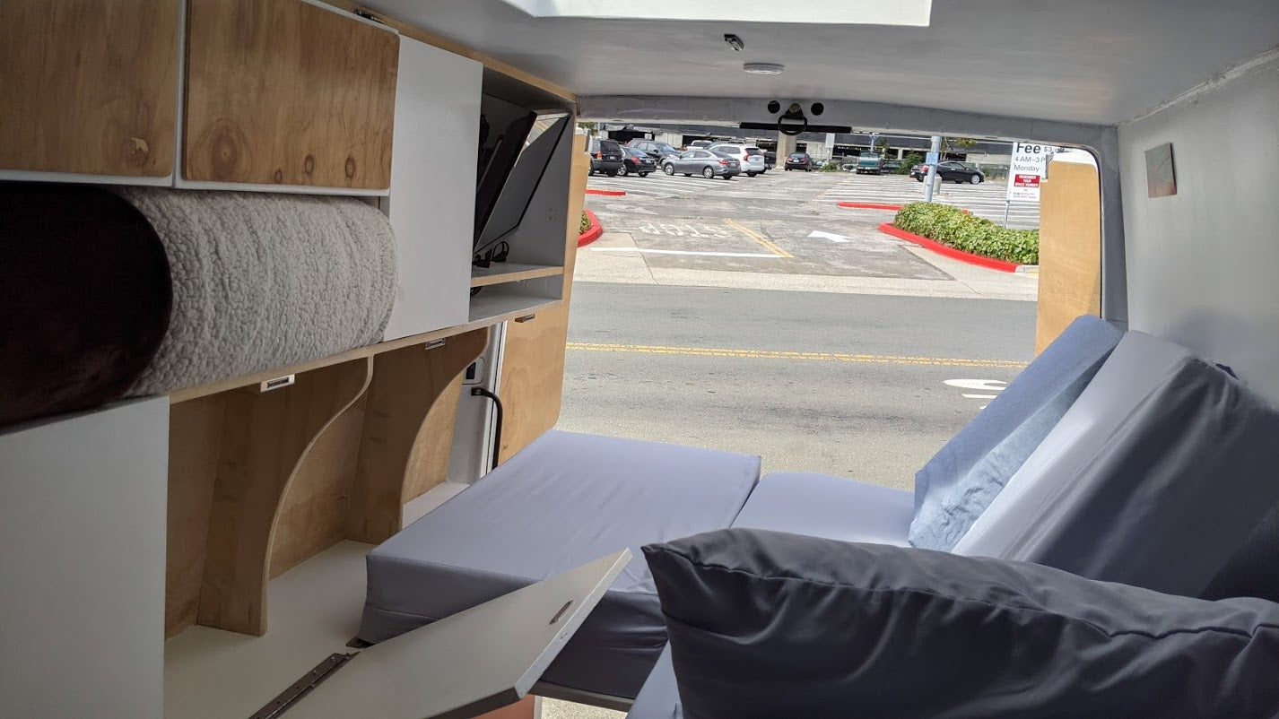 Inside an RV rental camper van: modular wood/white cabinetry, folded grey cushions forming a bed, looking out to a parking lot.