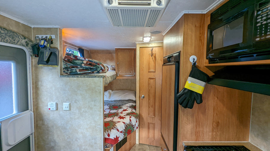 Inside an RV rental, showing compact living space with bunk beds, kitchen (microwave, fridge), and ceiling AC unit.