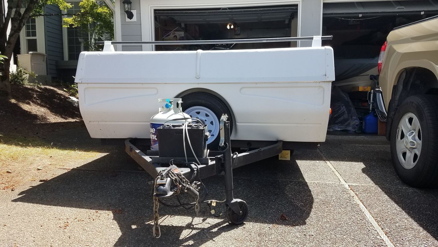 White utility trailer with propane tank and battery, parked on a driveway next to a truck. An ideal setup for an rv rental excursion.