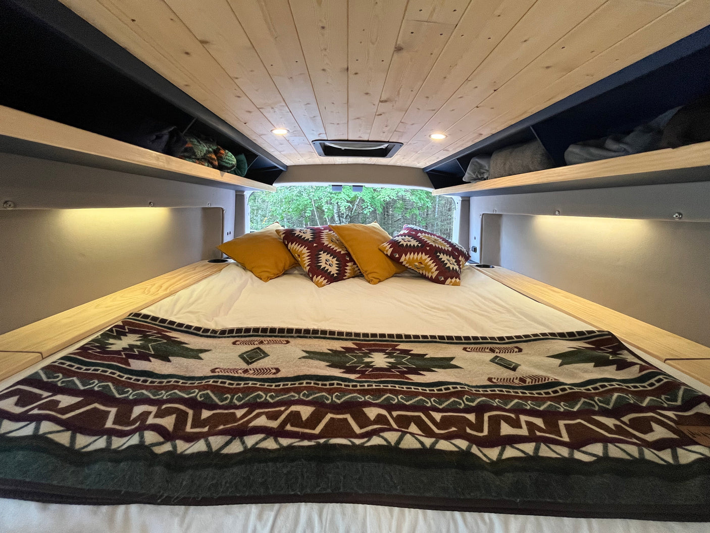 Interior of an **RV rental** featuring a cozy bed with patterned blanket, overhead storage, and nature view.