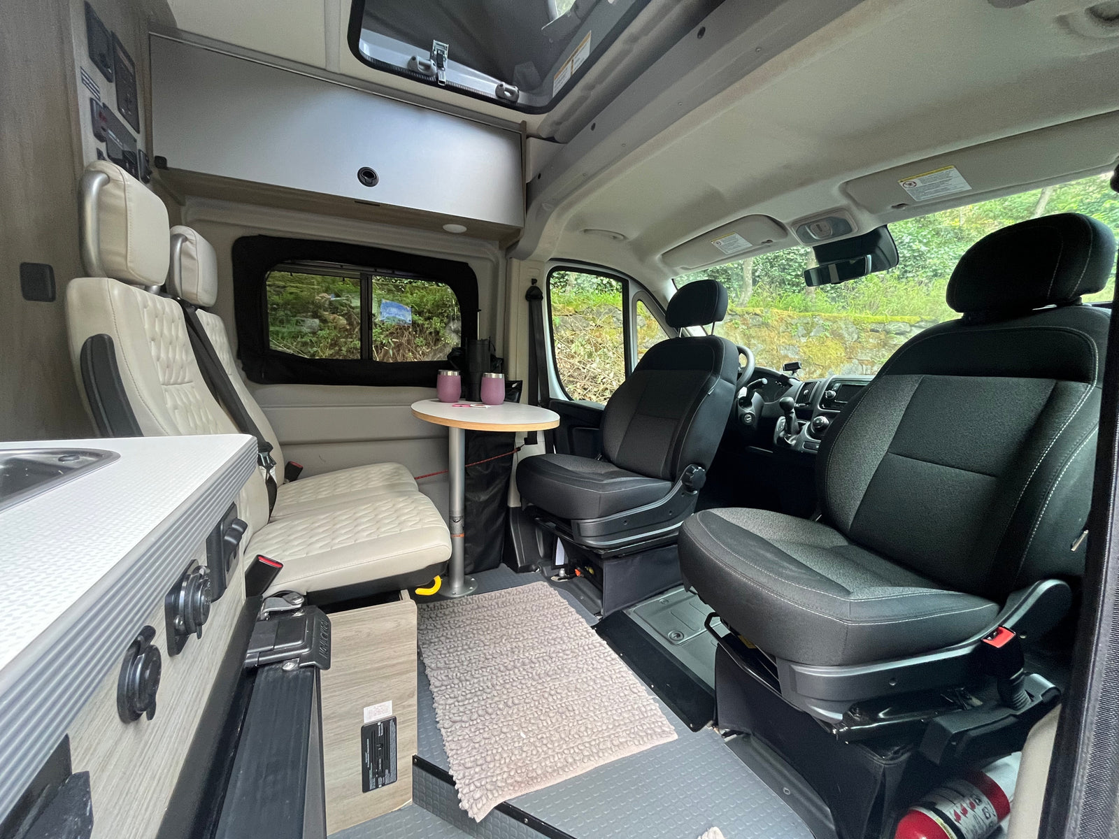 View inside an RV rental showing the living space. Features front swivel seats, a dining area with table & bench seats, and a compact kitchen.