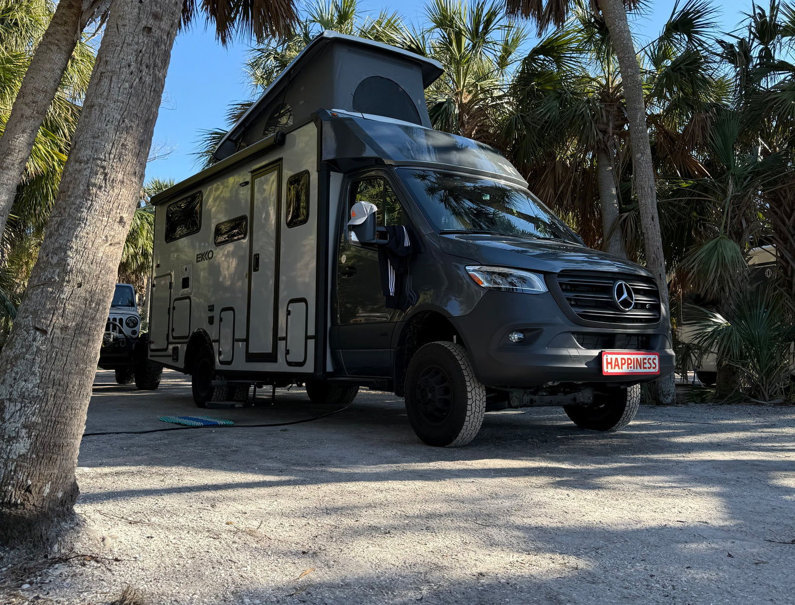 Gray EKKO RV rental (Mercedes camper van) with pop-top, parked on a shell lot under palm trees. Its plate reads HAPPINESS!