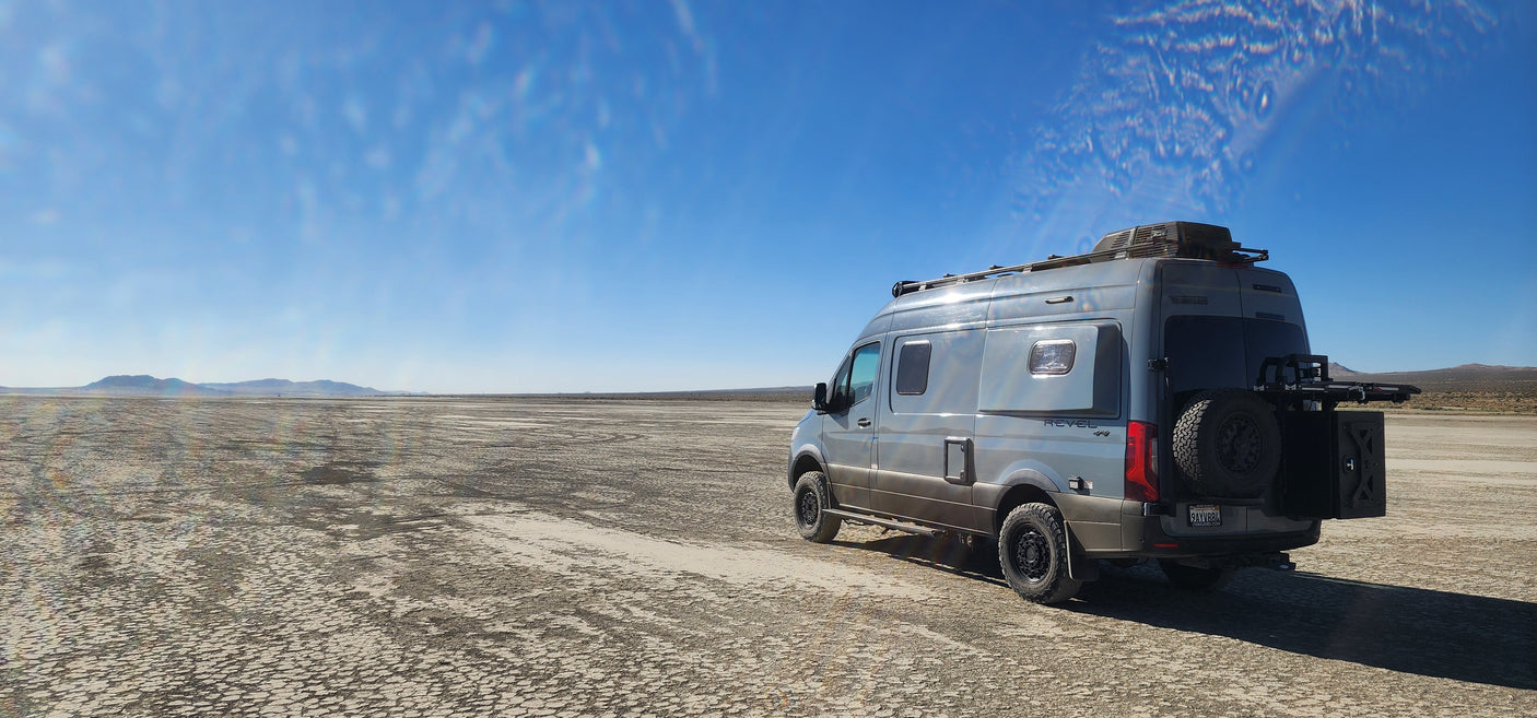 Grey Winnebago Revel RV rental on a vast, cracked dry lakebed under a clear blue sky, mountains afar.