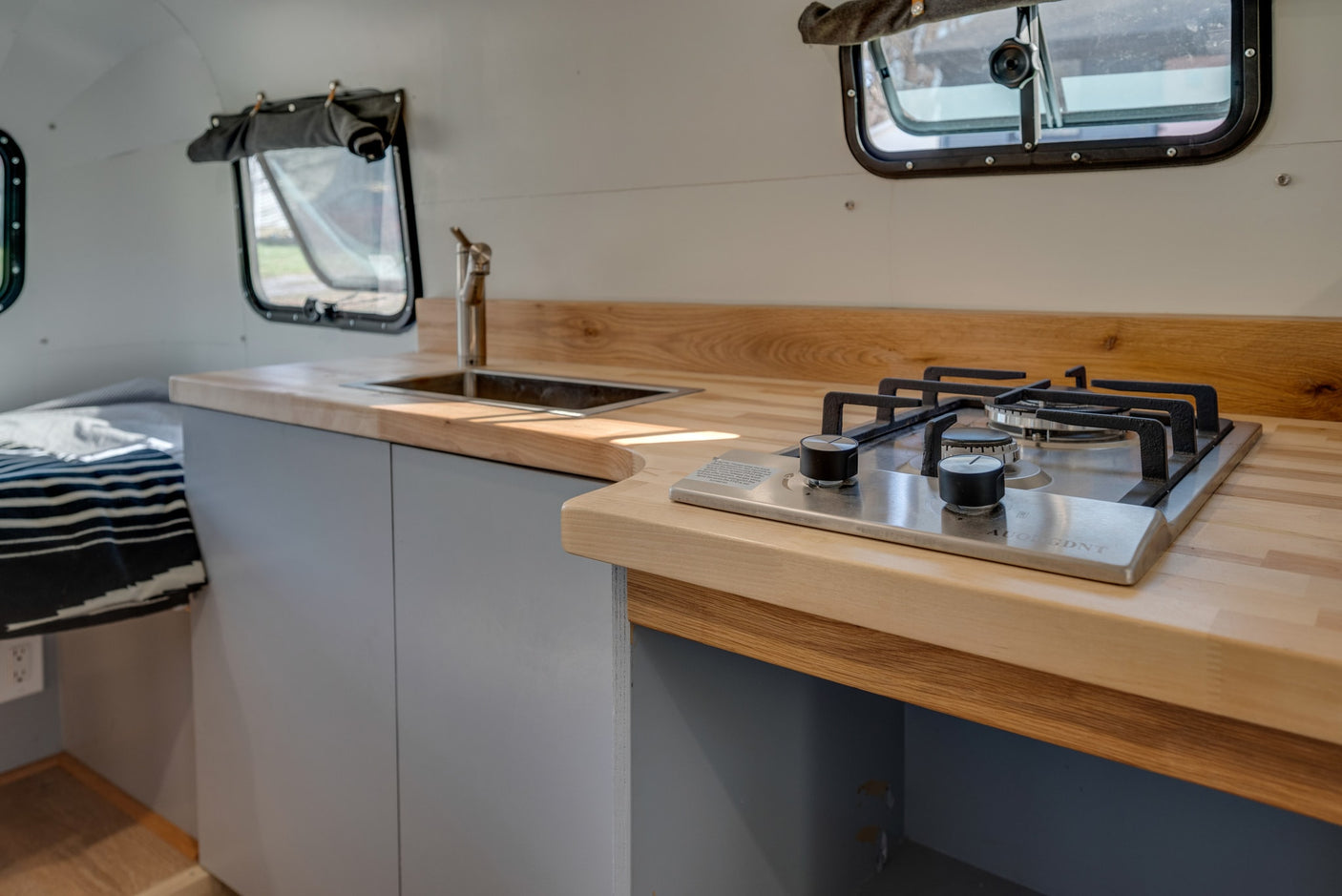 This RV rental interior features a kitchen with wood countertops, sink, and two-burner stove, adjacent to the bed.