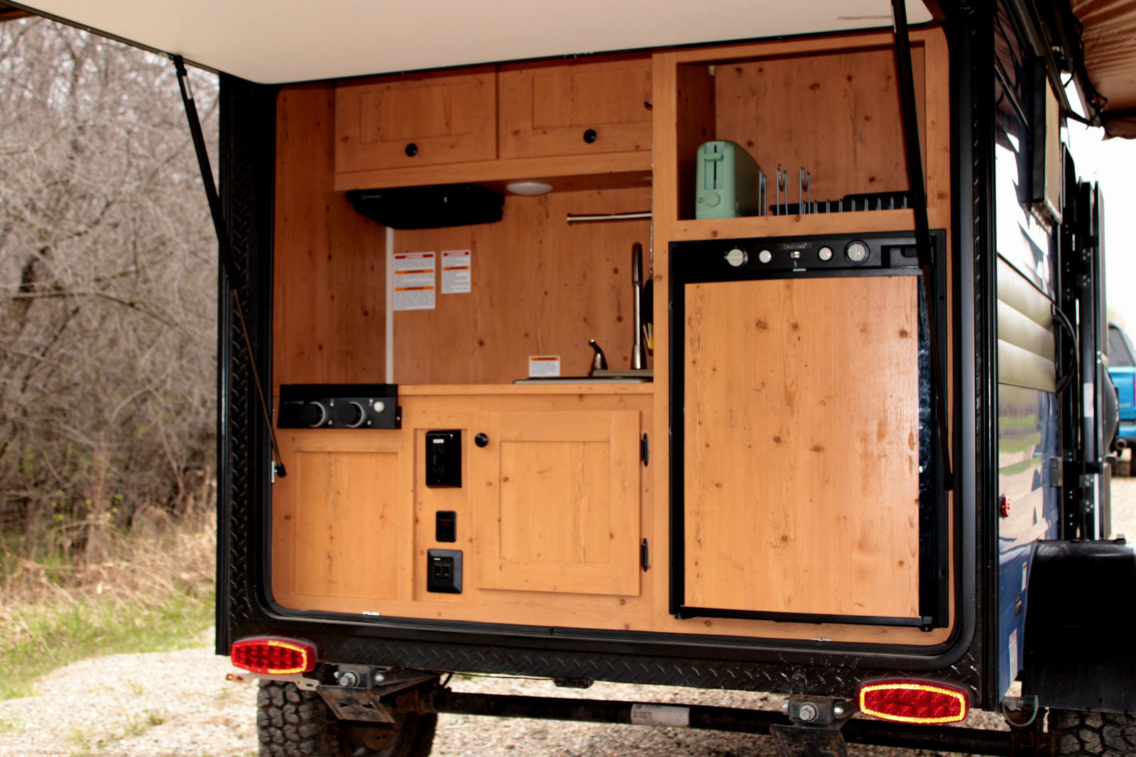 RV rental: Rear galley of a camper trailer featuring a wooden outdoor kitchen with a fridge, sink, and toaster, ready for adventure.