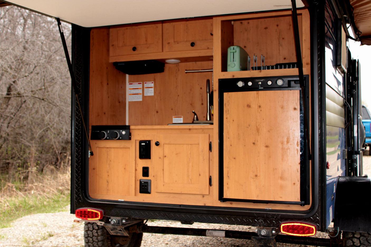 RV rental: Rear galley of a camper trailer featuring a wooden outdoor kitchen with a fridge, sink, and toaster, ready for adventure.