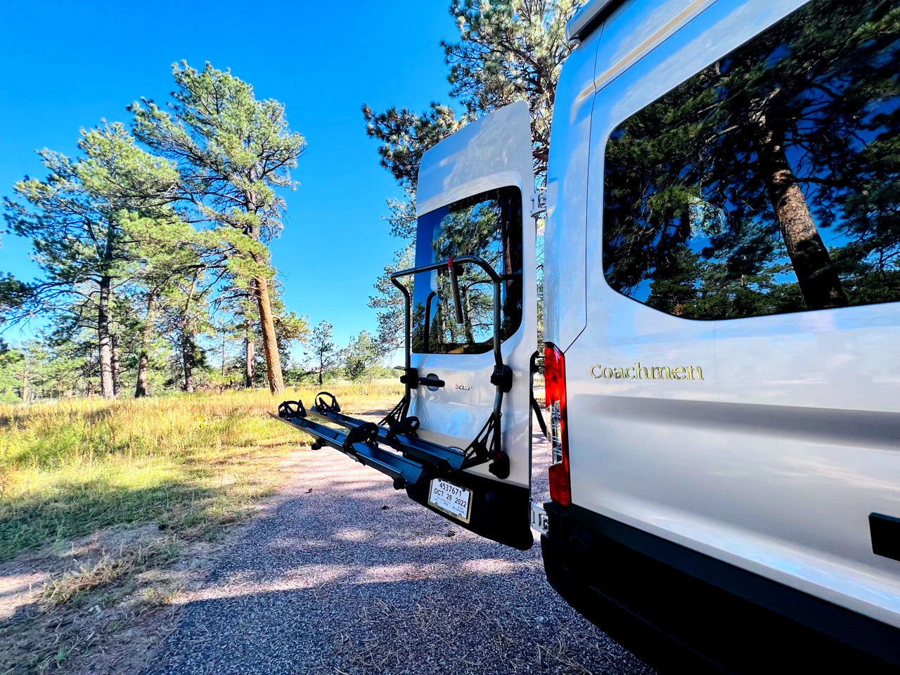 Rear view of a white Coachmen camper van RV rental with a bike rack, parked among pine trees for camping.