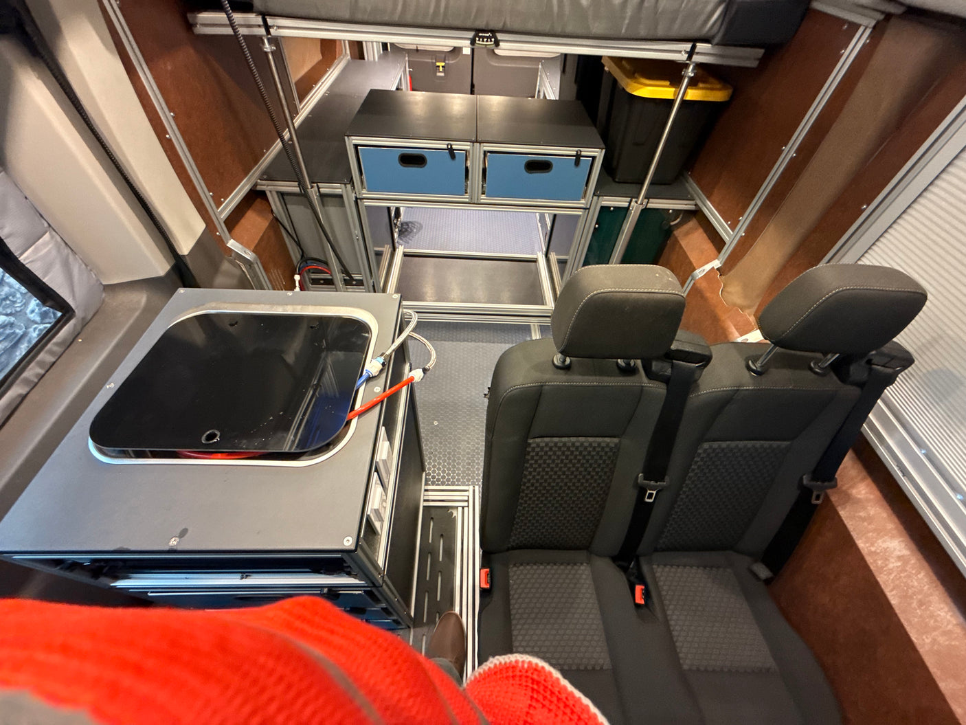 RV rental interior: Person's leg in foreground, rear seats, kitchenette, blue-drawer storage, and overhead bed.