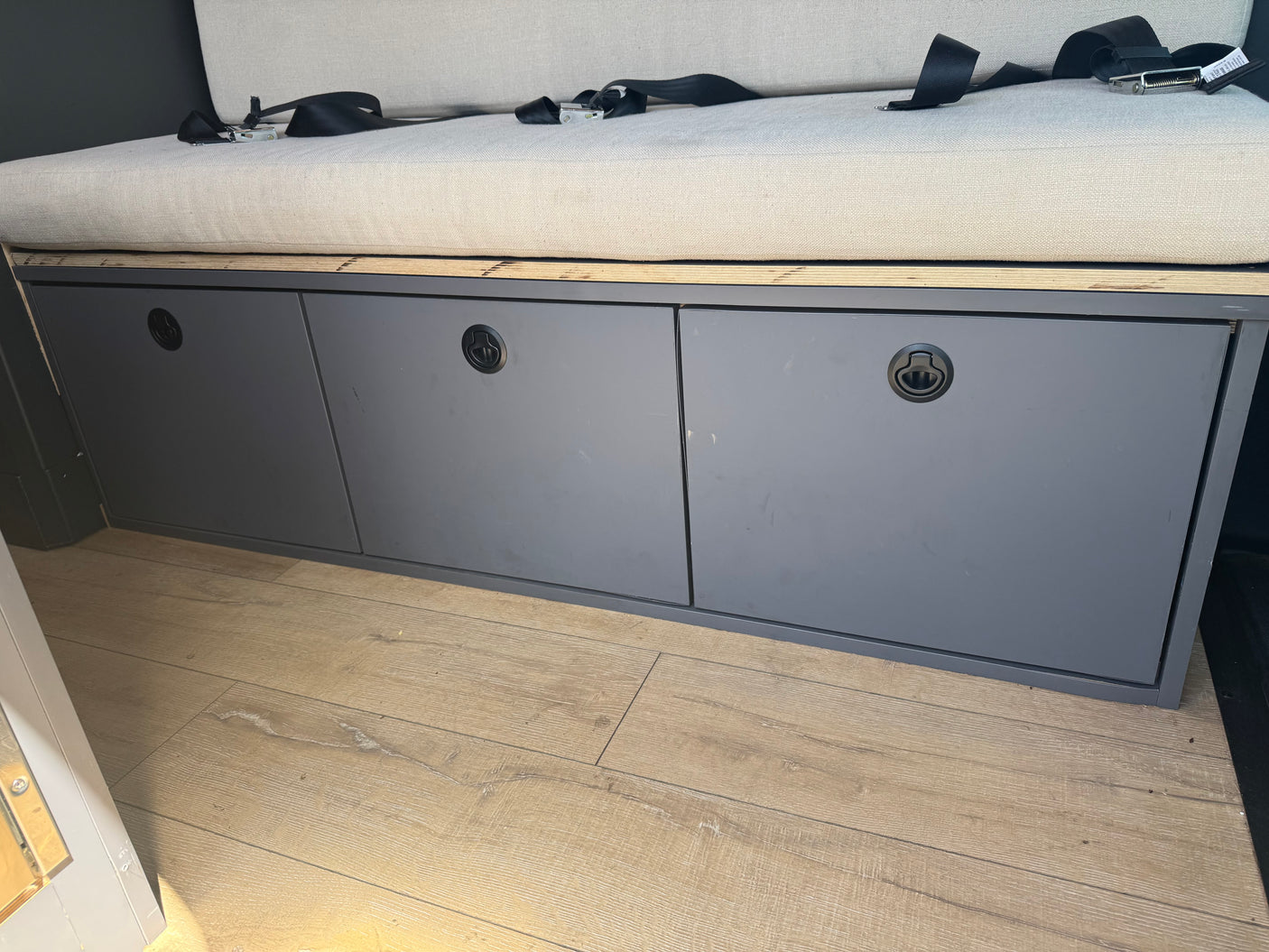 RV rental interior: Bench seat with safety belts above three dark gray storage cabinets and wood-look flooring.