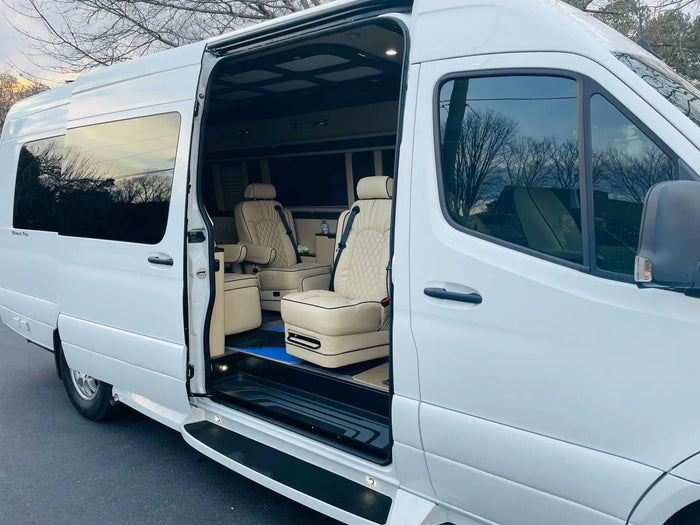 parked on asphalt with trees in the background. Ready for adventure.|Luxury white RV rental van with open side door revealing cream captain's chairs and opulent interior. Ideal for premium travel.|Spacious luxury van RV rental interior featuring multiple cream captain's chairs & modern amenities.|View of a high-end RV rental interior