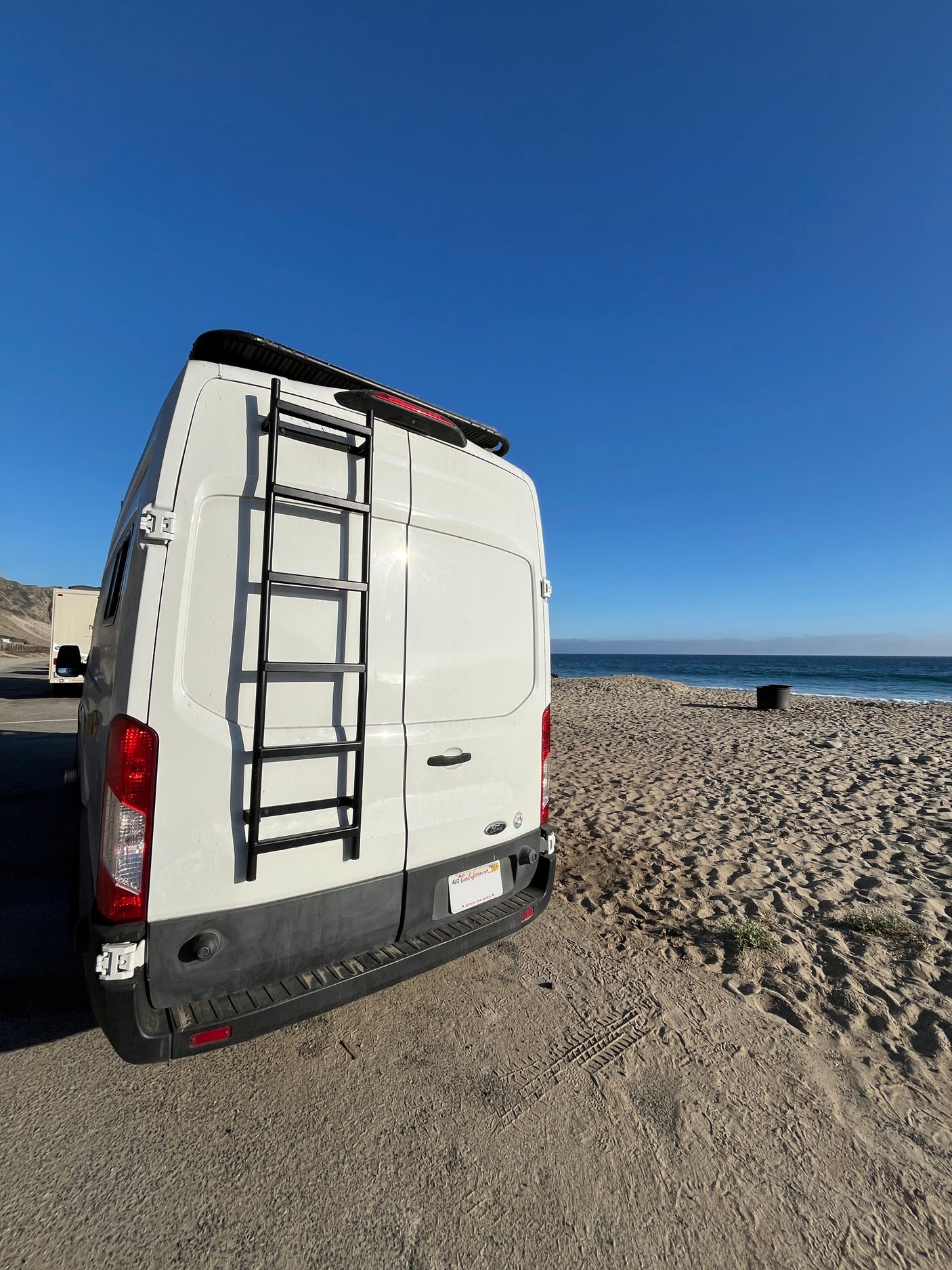 White RV rental van with a ladder, backed up to a sandy California beach. Clear blue ocean and sky complete the scenic view.