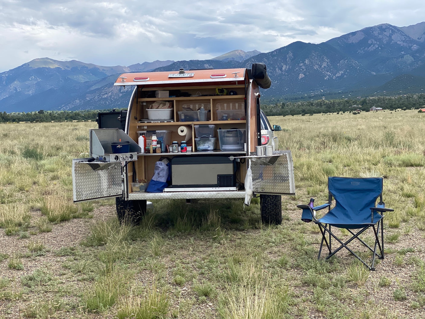 Perfect RV rental for outdoor cooking! This camper's kitchen is open in a scenic mountain field with a chair.