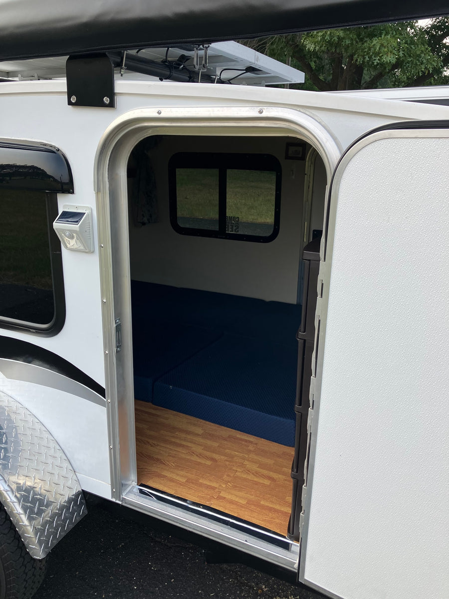 Compact white travel trailer RV rental interior with blue mattress, wood floor, solar light. Awning & solar panel visible.