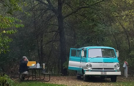 Man at a picnic table next to a vintage teal & white Dodge RV rental van in a wooded campsite.