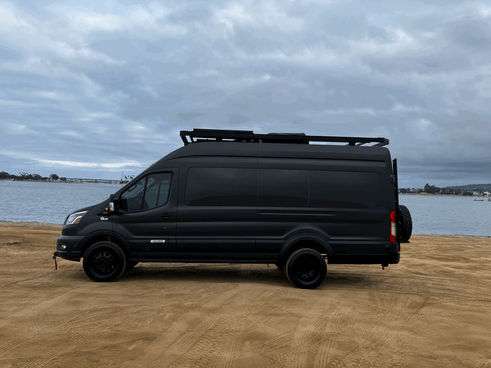 Black Vandoit camper van parked on sandy beach by water. Your next RV rental adventure awaits!