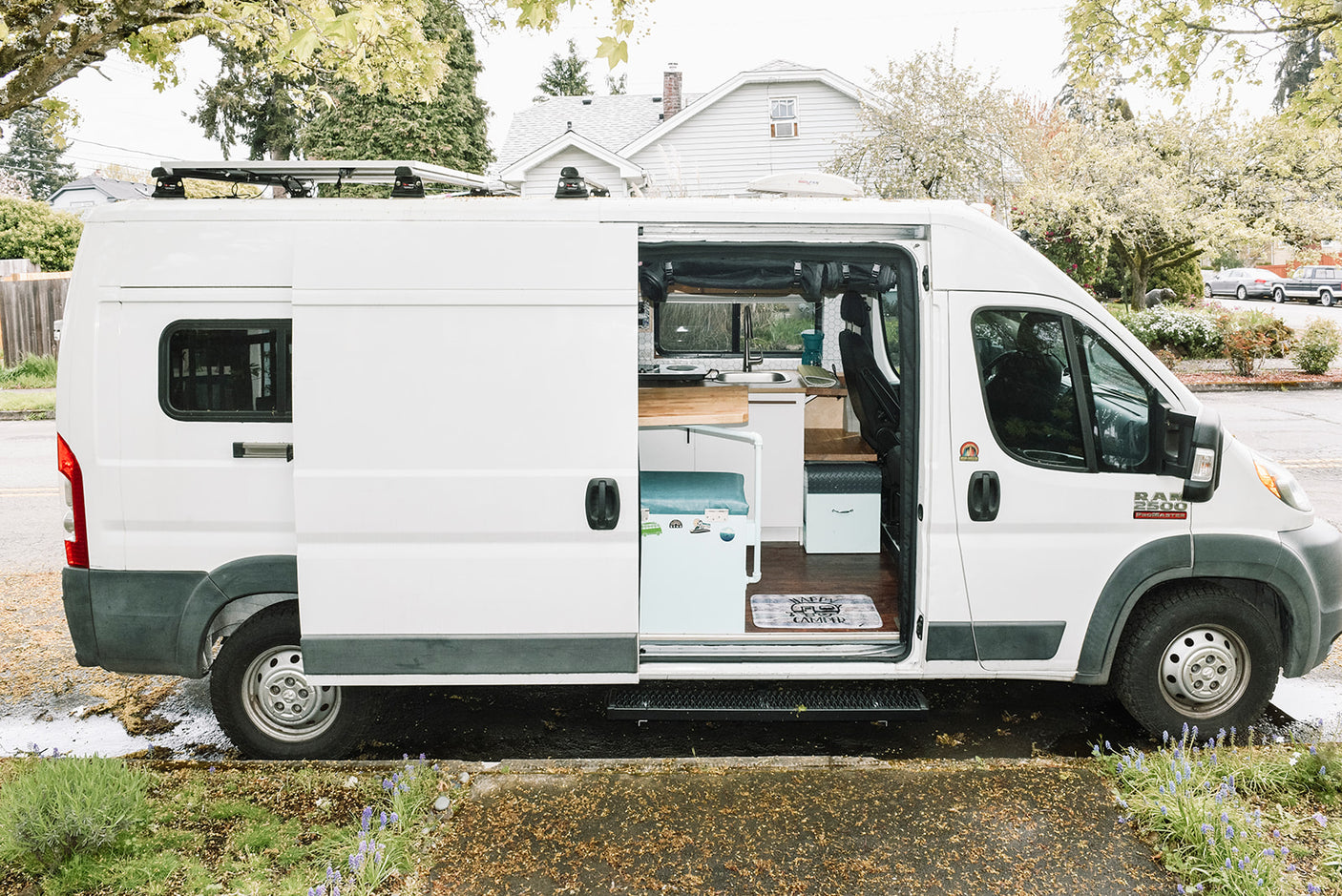 This white Ram ProMaster converted camper van, ideal for rv rental, has its side door open, revealing a compact kitchen and living area inside.
