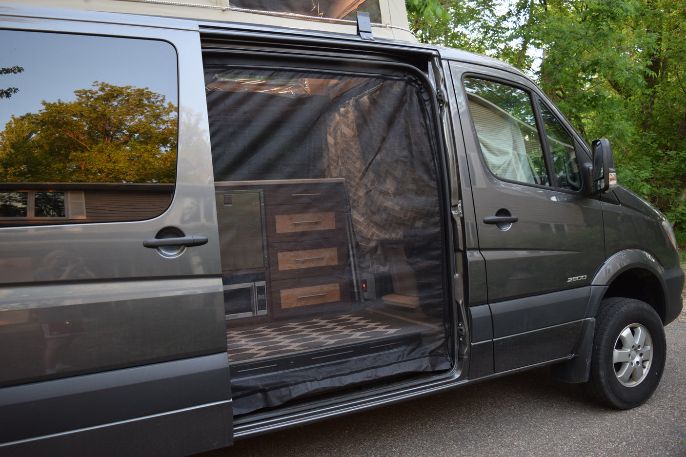 Explore this RV rental's cozy interior: a grey van with its side door open, featuring custom cabinets, microwave, and a bug screen.