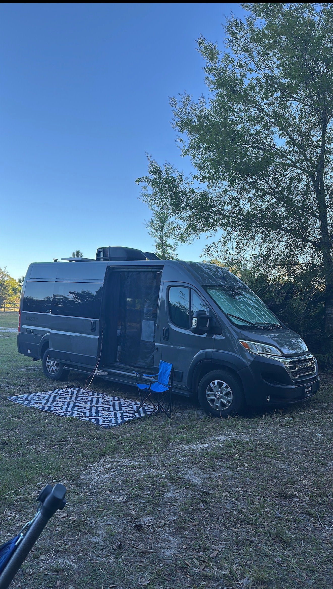 Our grey RV rental parked in a grassy spot. The campervan's side door is open, with a rug and blue chair set up for outdoor relaxation.