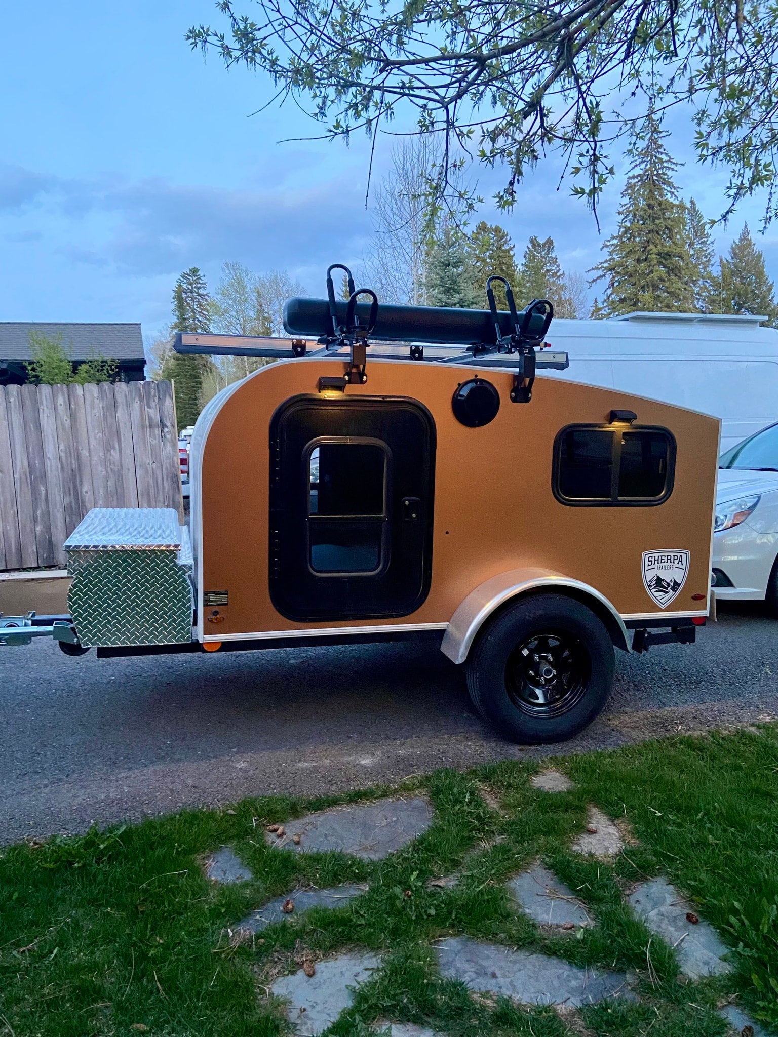 A tan Sherpa teardrop trailer, a compact RV rental with a roof rack and front storage box, parked on pavement. Ideal for travel.