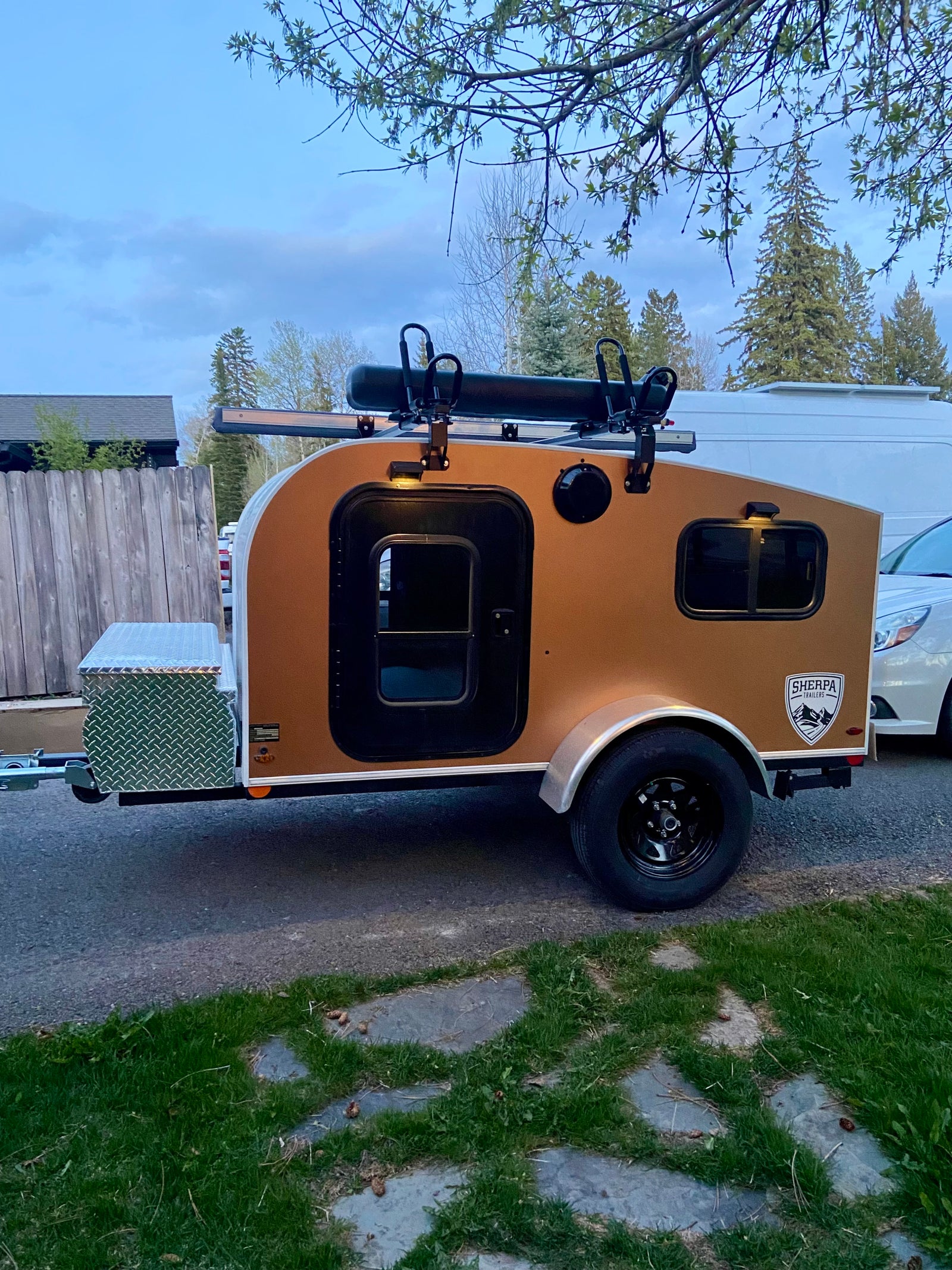 A tan Sherpa teardrop trailer, a compact RV rental with a roof rack and front storage box, parked on pavement. Ideal for travel.