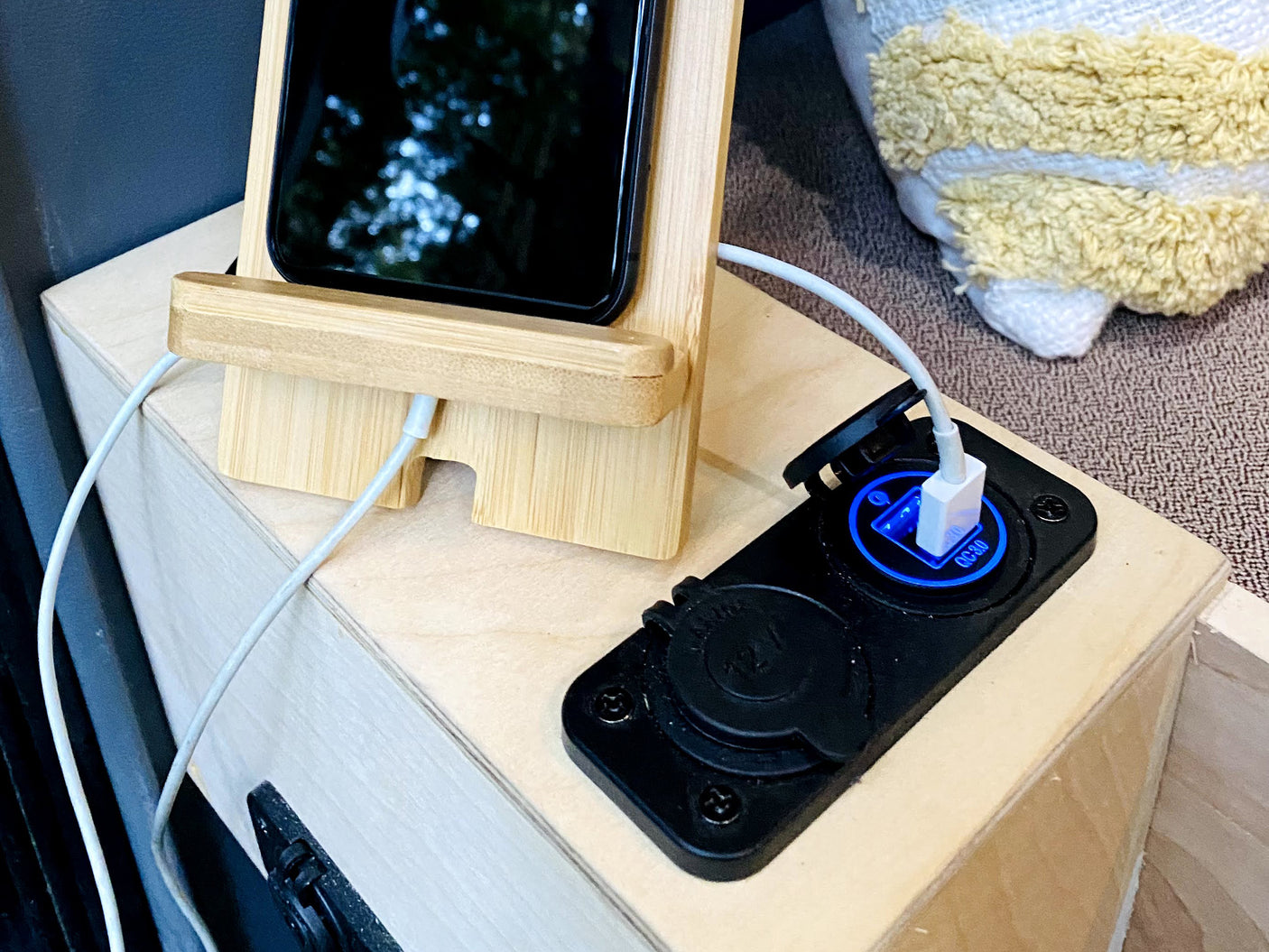 An RV rental interior: a smartphone on a bamboo stand charges via a blue-lit 12V USB port on a light wood surface.