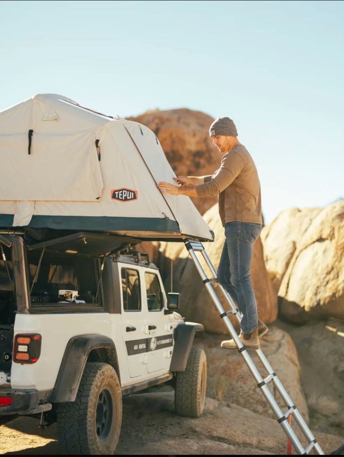 and person on ladder. Ideal RV rental.|Woman enjoying a meal from an RV rental Jeep Gladiator's camping kitchen in a stunning desert landscape.|Man sets up a rooftop tent on a white Jeep RV rental with a ladder. Ready for desert camping adventures!|Woman smiles from her Tepui rooftop tent atop a Jeep Gladiator