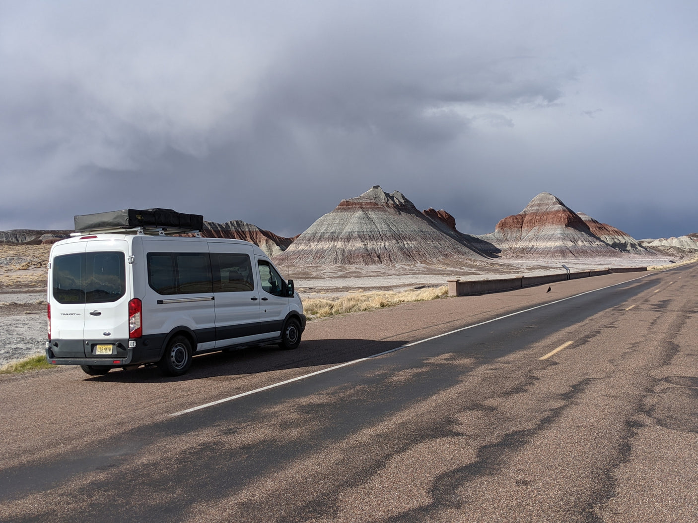White Ford Transit RV rental parked at a stunning desert overlook with layered mountains & dramatic clouds.|RV rental van with rooftop tent set up at a peaceful campsite