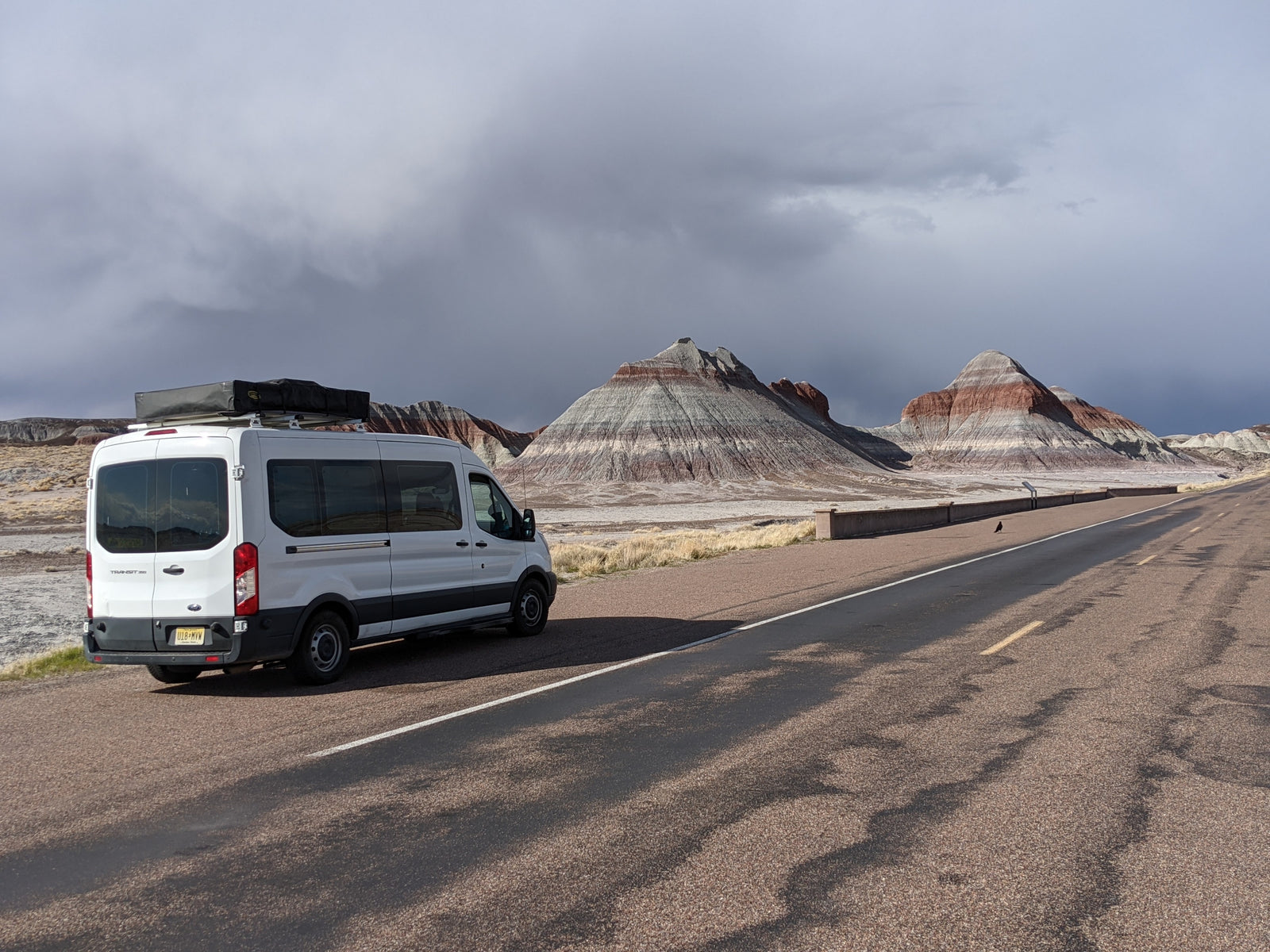 White Ford Transit RV rental parked at a stunning desert overlook with layered mountains & dramatic clouds.|RV rental van with rooftop tent set up at a peaceful campsite
