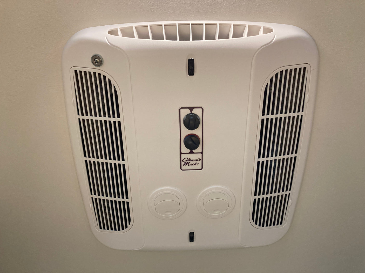 This white, ceiling-mounted Coleman Mach AC unit, common in RV rentals, offers climate control via its knobs and adjustable vents.