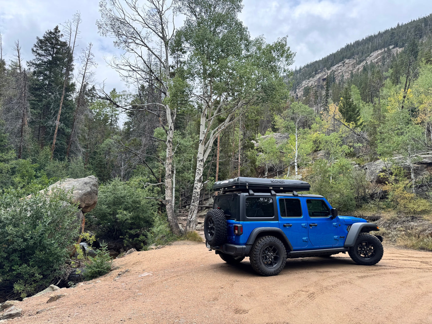 Blue Jeep with a rooftop tent parked on a forest dirt road, surrounded by trees. A great rv rental for mountain adventures.
