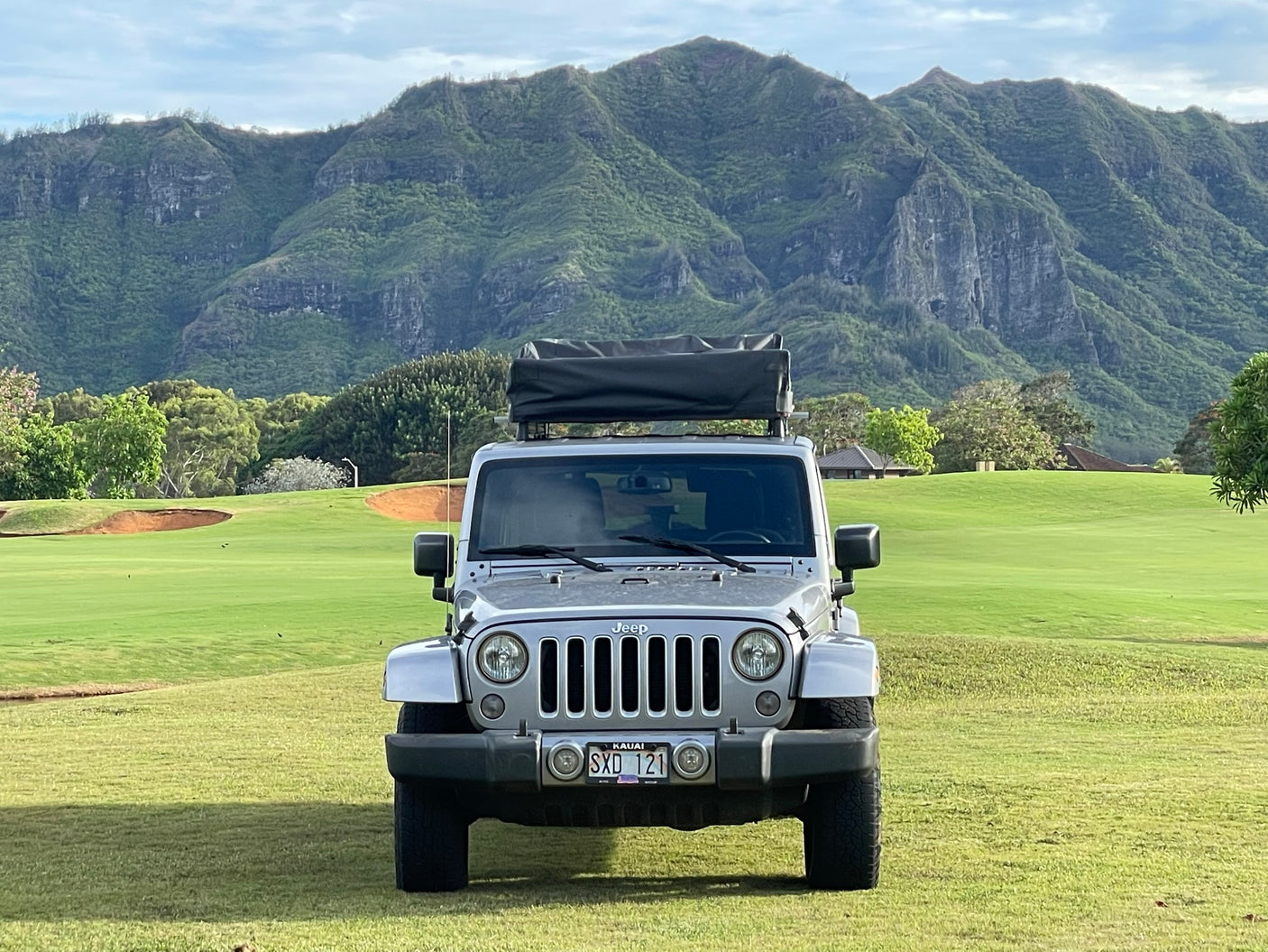 Parked on Kauai, a silver Jeep with a rooftop tent faces lush mountains. Perfect for your outdoor RV rental experience.