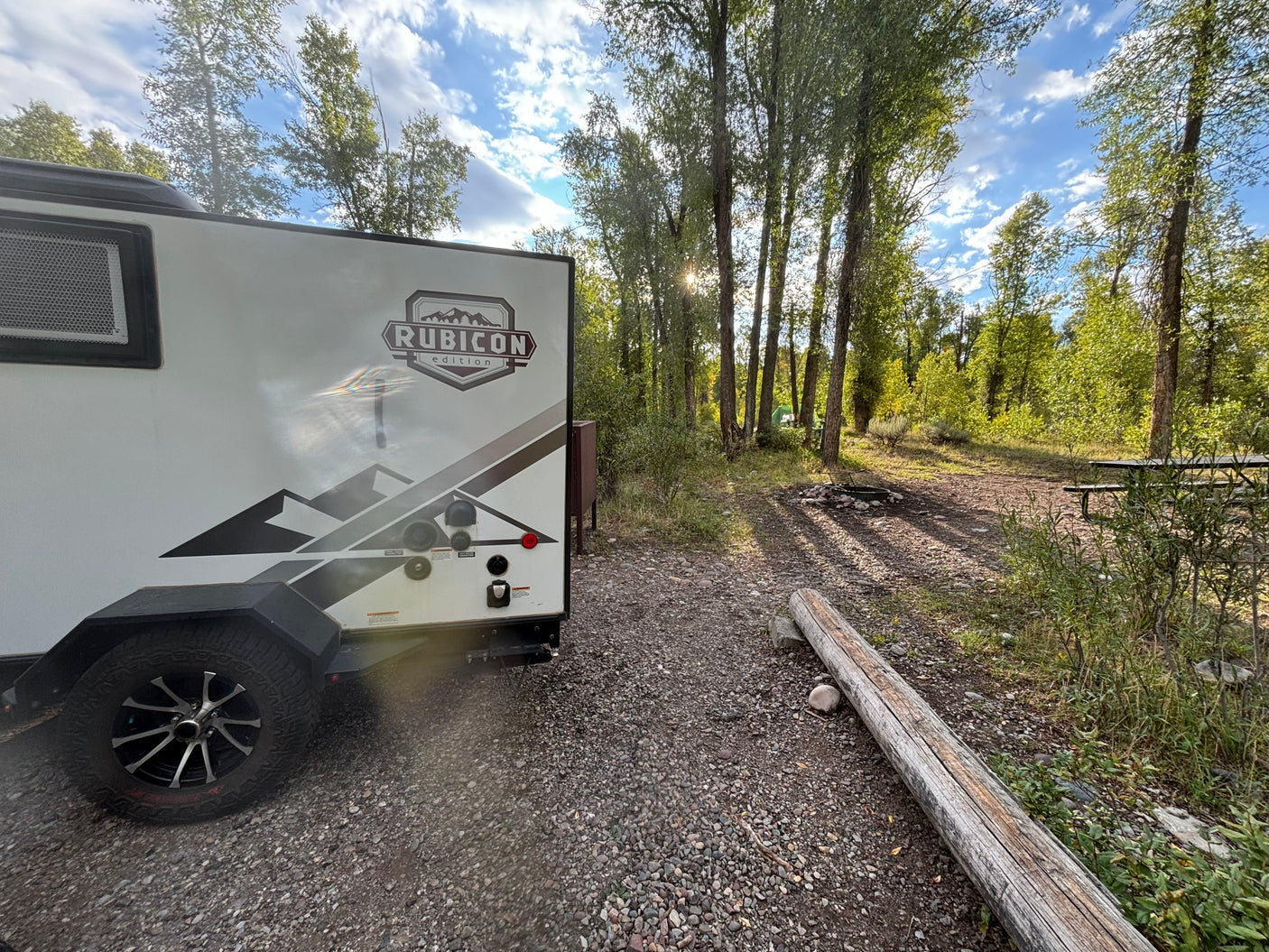 Rubicon Edition RV rental at a sunny gravel campsite with a fire pit, picnic table, and tall trees.