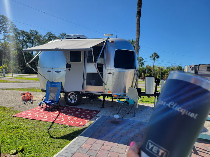 RV rental setup: A shiny Airstream travel trailer with awning extended, chairs, and rug, ready for leisure in a sunny campground.
