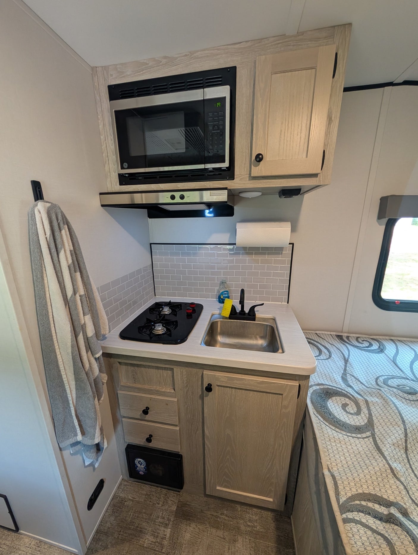 Modern rv rental kitchen with microwave, stove, sink, and light-toned cabinets. Towel and bed visible for context.