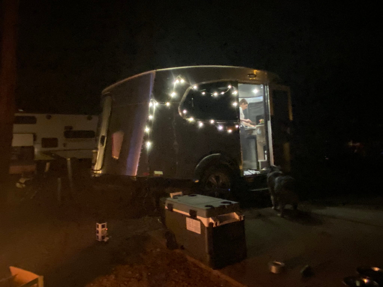 Nighttime RV rental scene: An Airstream-style camper lit with string lights, a person inside, and a dog outside.