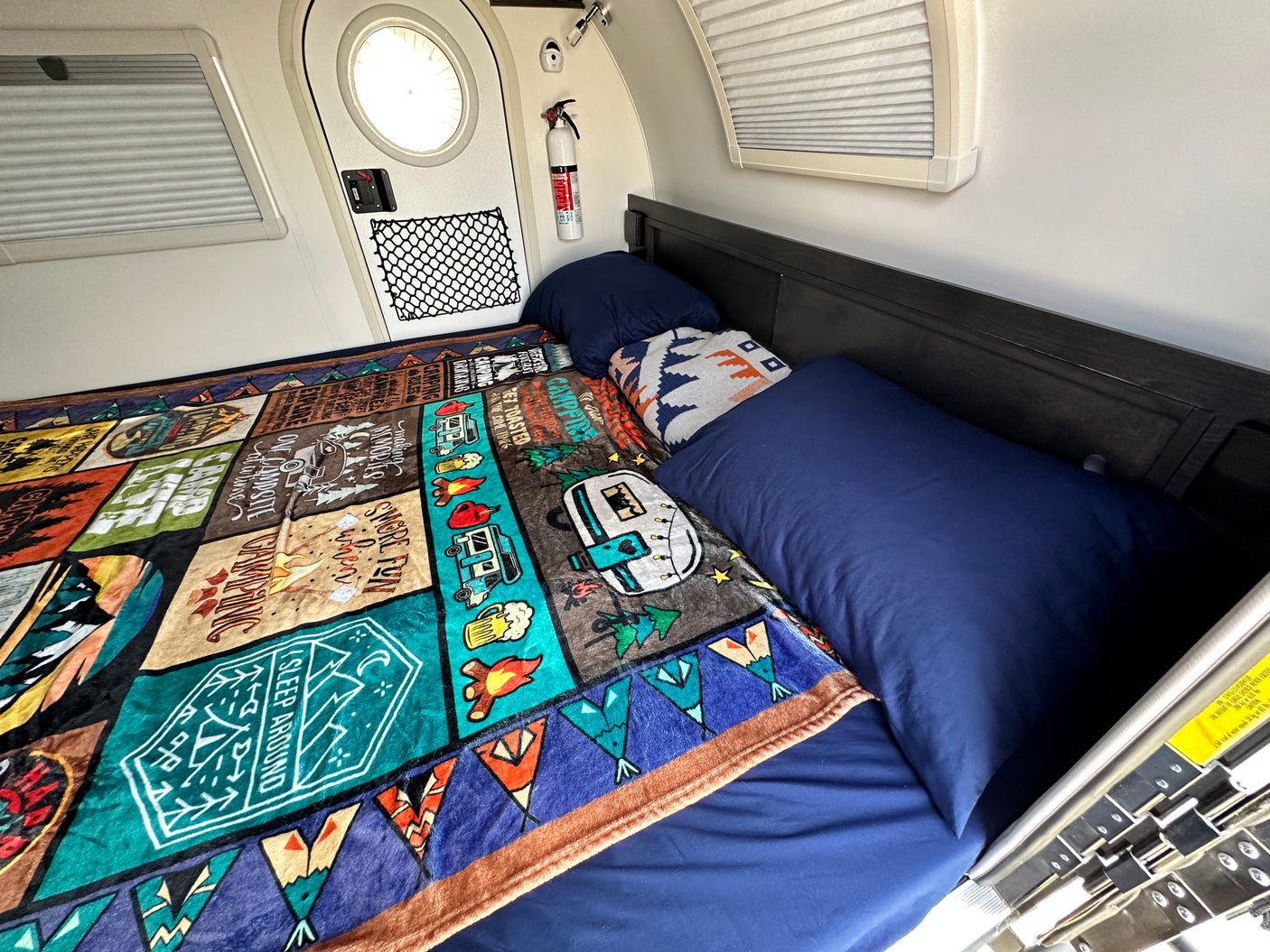 Interior of an RV rental showing a bed with blue linens & a colorful camping blanket, fire extinguisher, and windows.