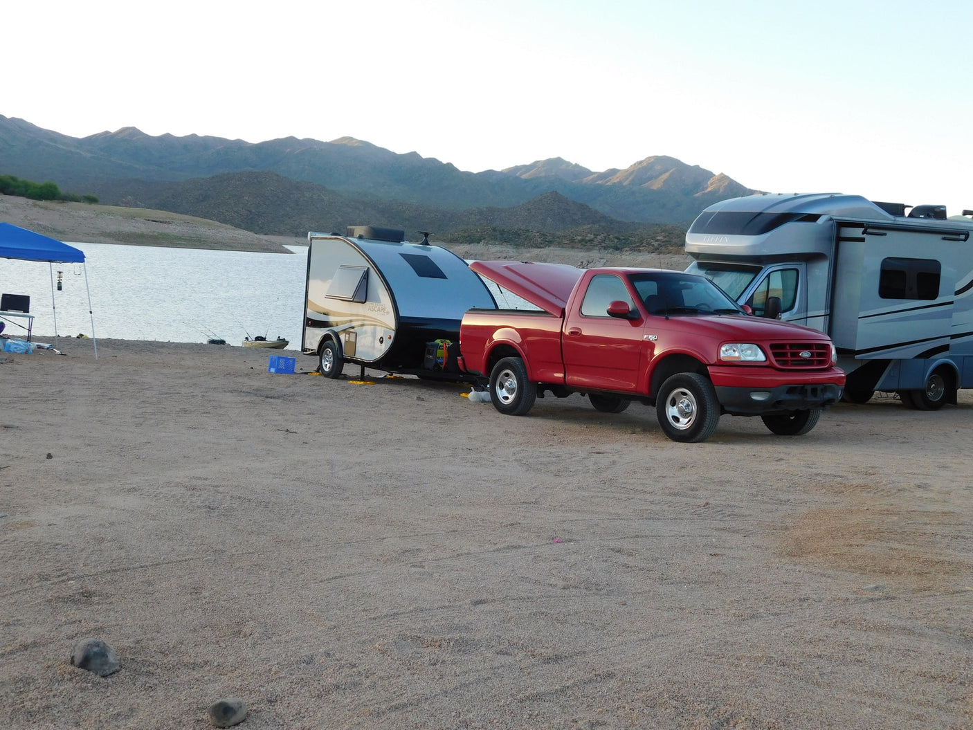 RV rental vehicles: Red pickup towing a Grand Ascape camper, and a Tiffin motorhome, parked by a lake and mountains.