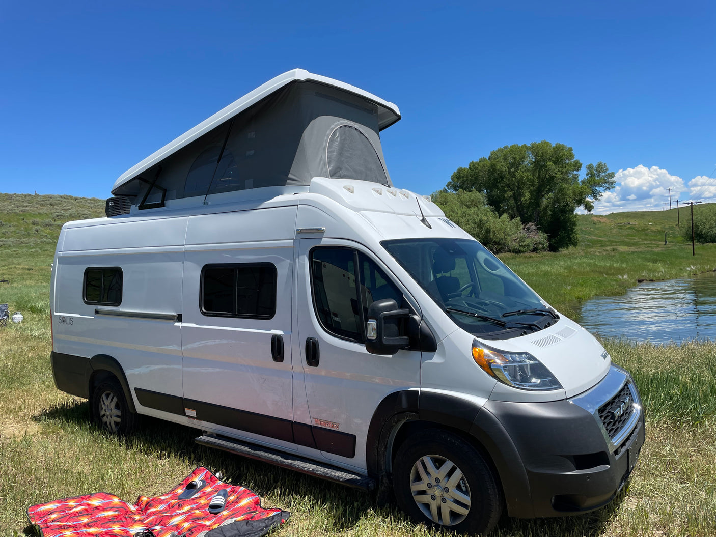 Explore nature with this Winnebago Solis pop-top camper van rv rental, parked by water under a clear blue sky.