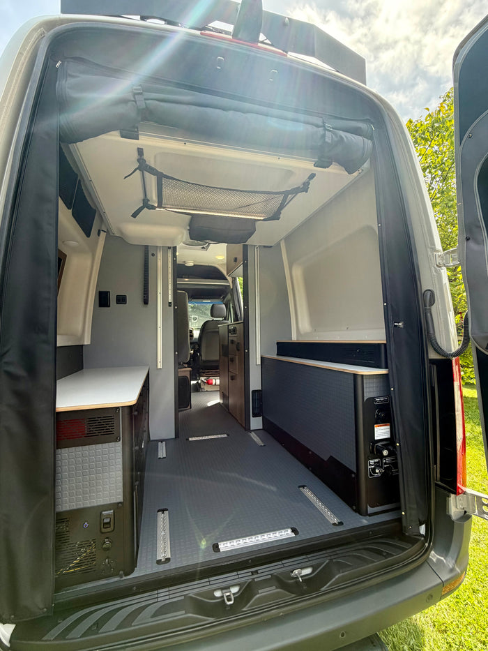 Rear view of an RV rental camper van interior: compact kitchen, utility panel, flexible floor tracks, and overhead storage.