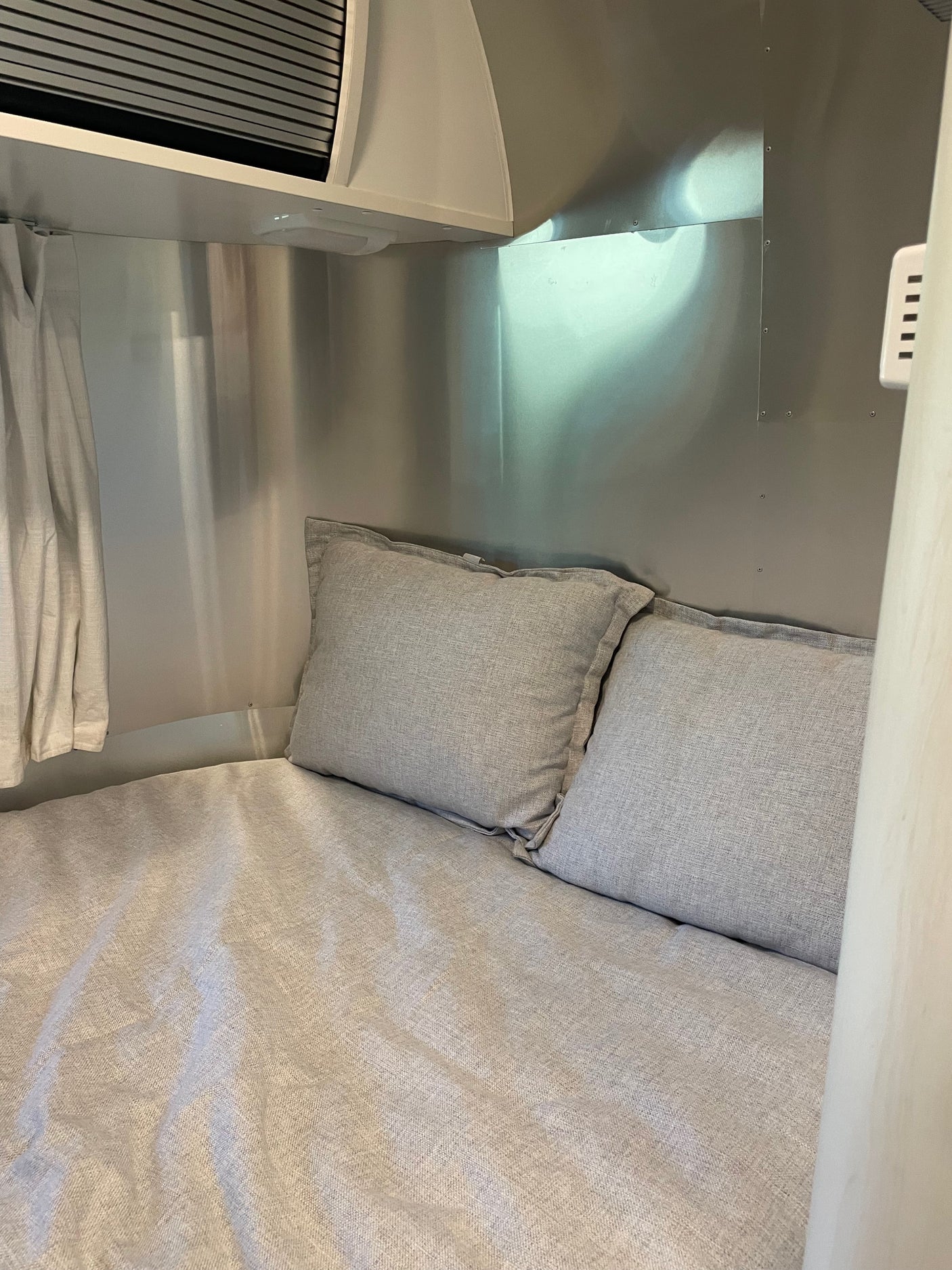 Cozy RV rental bed with light gray linen bedding and pillows, set within a sleek metallic travel trailer interior.