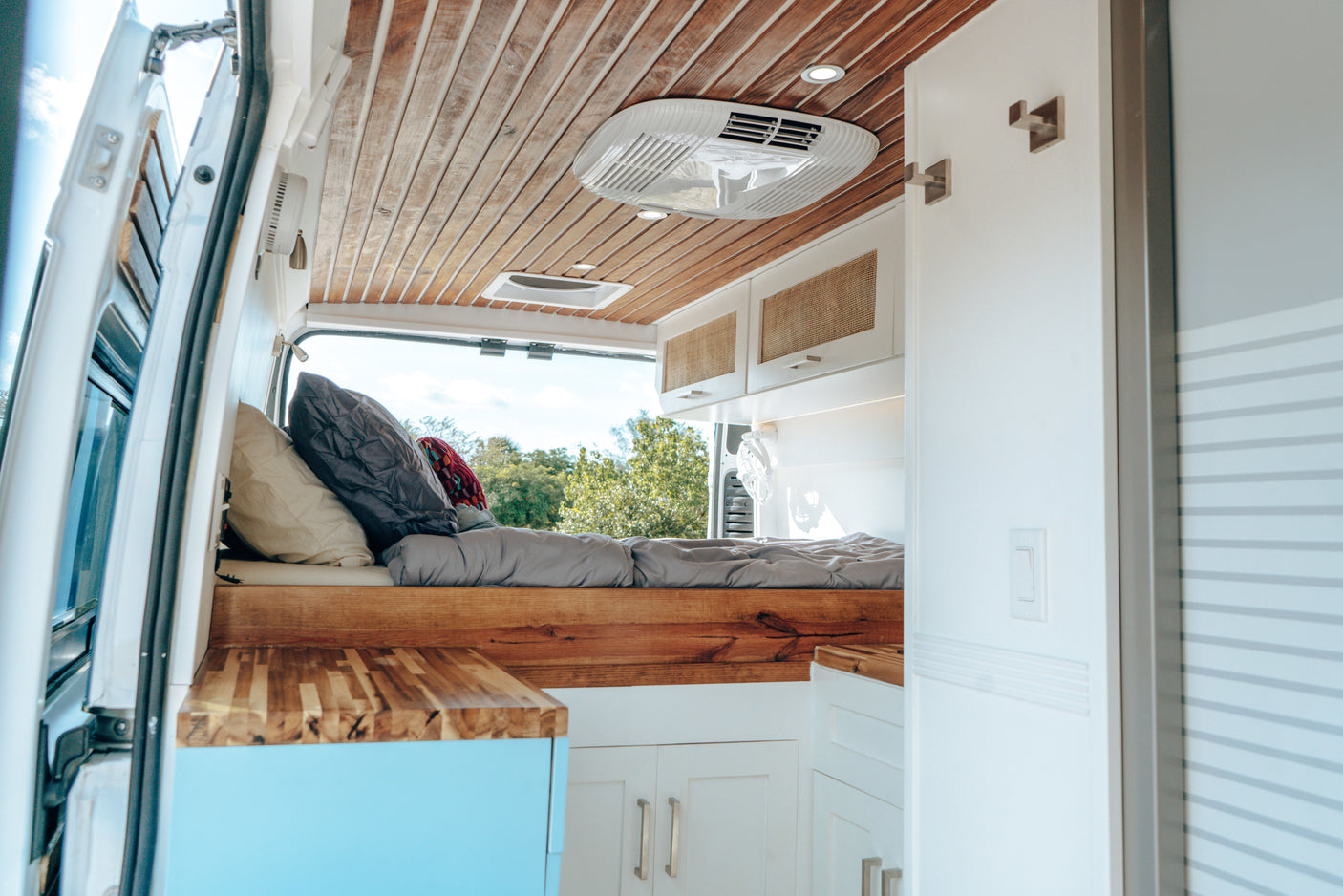 Inside a bright RV rental: view of the comfy bed, wooden ceiling, and compact kitchen/living area with AC unit.