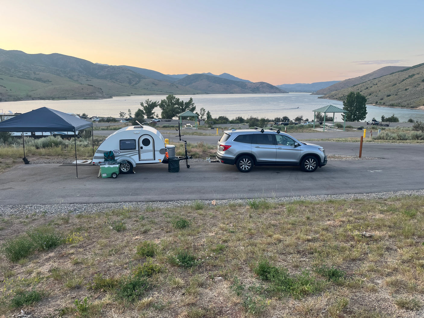 RV rental: Silver SUV towing a teardrop trailer set up at a scenic lakeside campsite with mountains under a soft sky.
