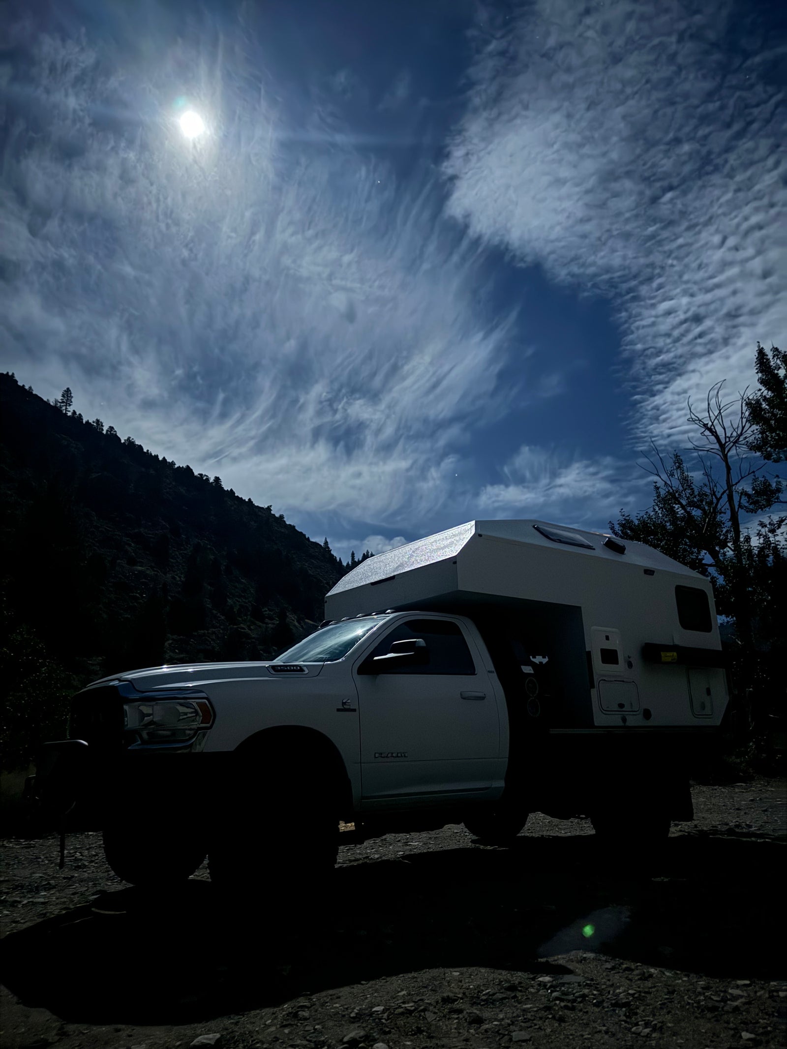 An overlanding truck camper RV rental parked beneath a bright sun and swirling clouds in a dark mountain landscape.