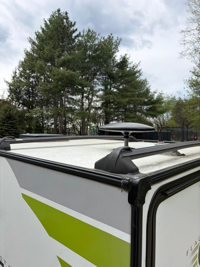 RV rental's roof with satellite dish & racks, green-striped side visible. Pine trees in background under cloudy sky.