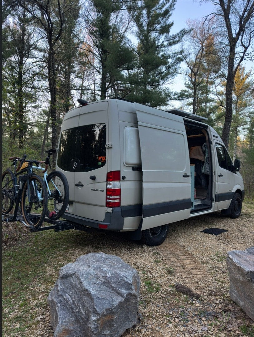 This beige rv rental camper van with bikes is parked in a natural, wooded setting, doors open for an outdoor adventure. — photo 7