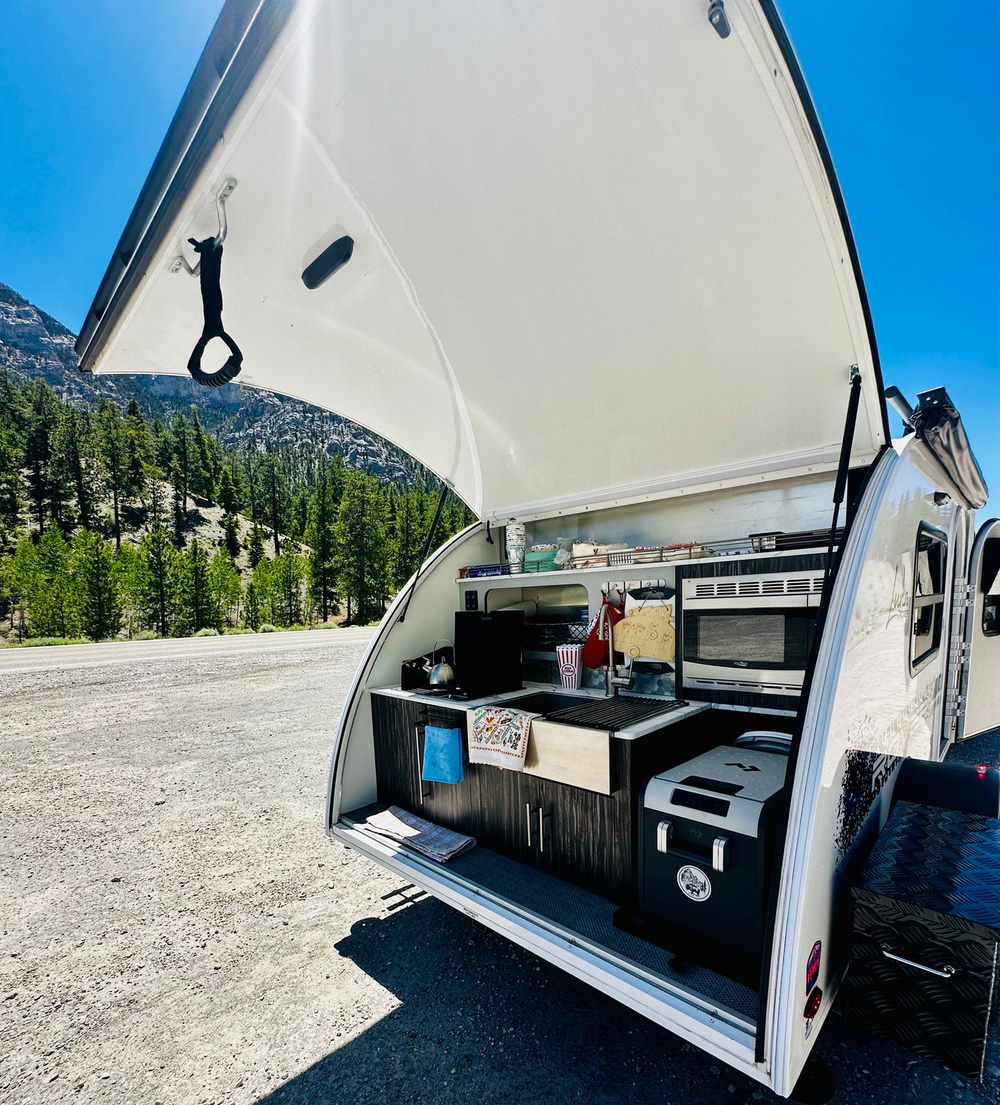 RV rental outdoor kitchen with microwave, sink, and cooler, showcasing mountain and forest scenery.