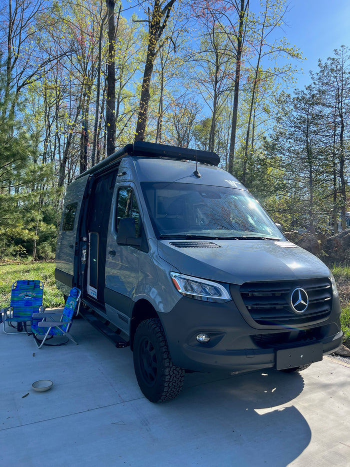 Grey Mercedes Sprinter RV rental camper van parked with open door, chairs, and fresh spring trees in a sunny outdoor setting.