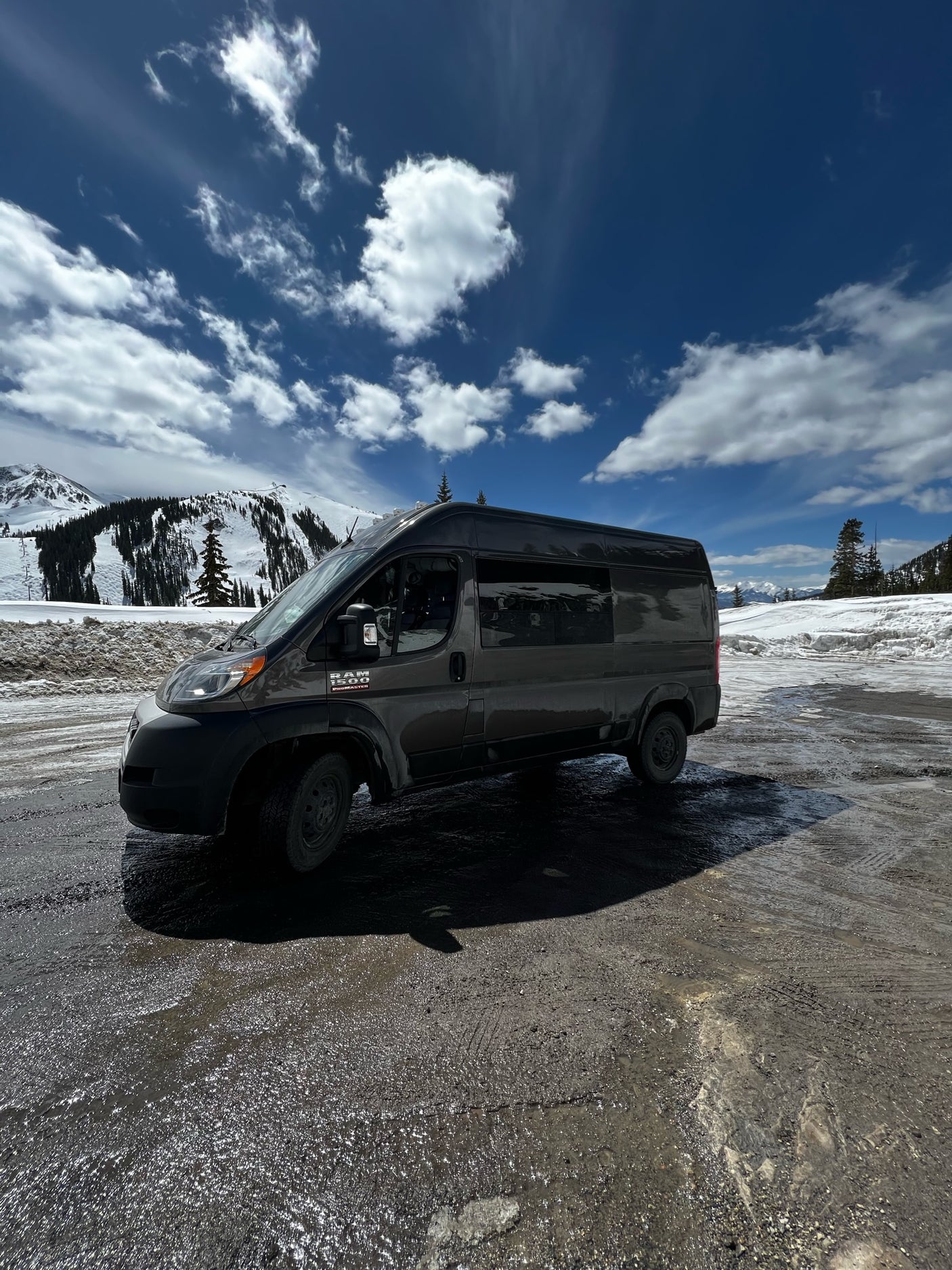 Dark grey Ram ProMaster RV rental parked on a wet mountain lot, snowy peaks & blue sky in the background. Ready for adventure!